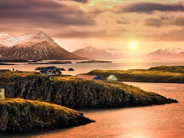 Coastal village with colorful houses on rocky islands, snow-capped mountains in the background, under a warm sunset sky.