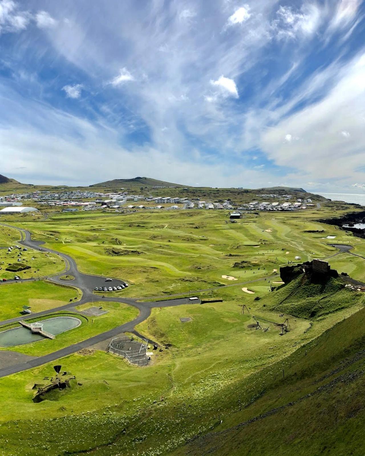 High-angle view of a sprawling green golf course and a coastal town under a blue sky with wispy clouds.