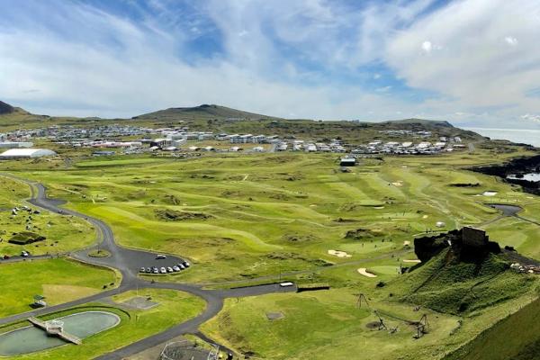 High-angle view of a sprawling green golf course and a coastal town under a blue sky with wispy clouds.