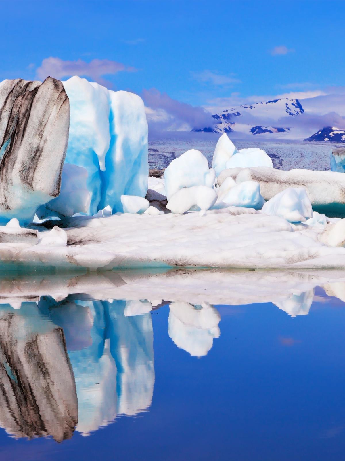 Icebergs, some with dark streaks, float in a calm blue glacial lagoon, reflecting in the water with snowy mountains in the background.