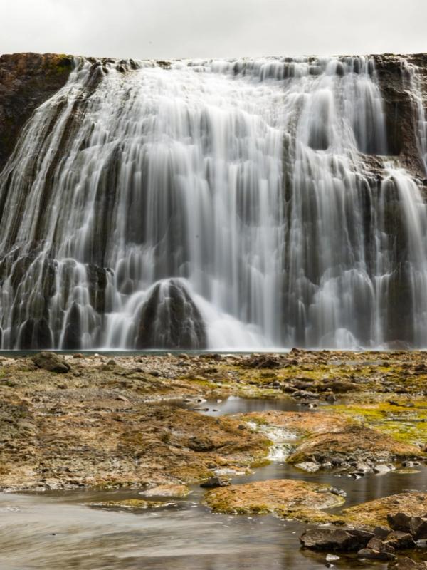 A wide waterfall cascades over rocky cliffs into a rocky foreground with green patches.