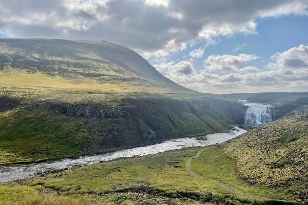 A wide river flows through a green, hilly valley towards a large waterfall in the distance under a cloudy sky.