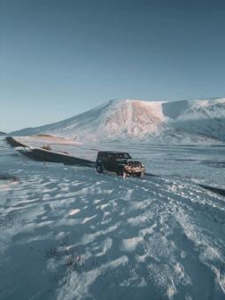 Un Jeep Wrangler negro aparcado en un arcén nevado junto a una carretera, con una gran montaña cubierta de nieve bajo un cielo azul despejado.