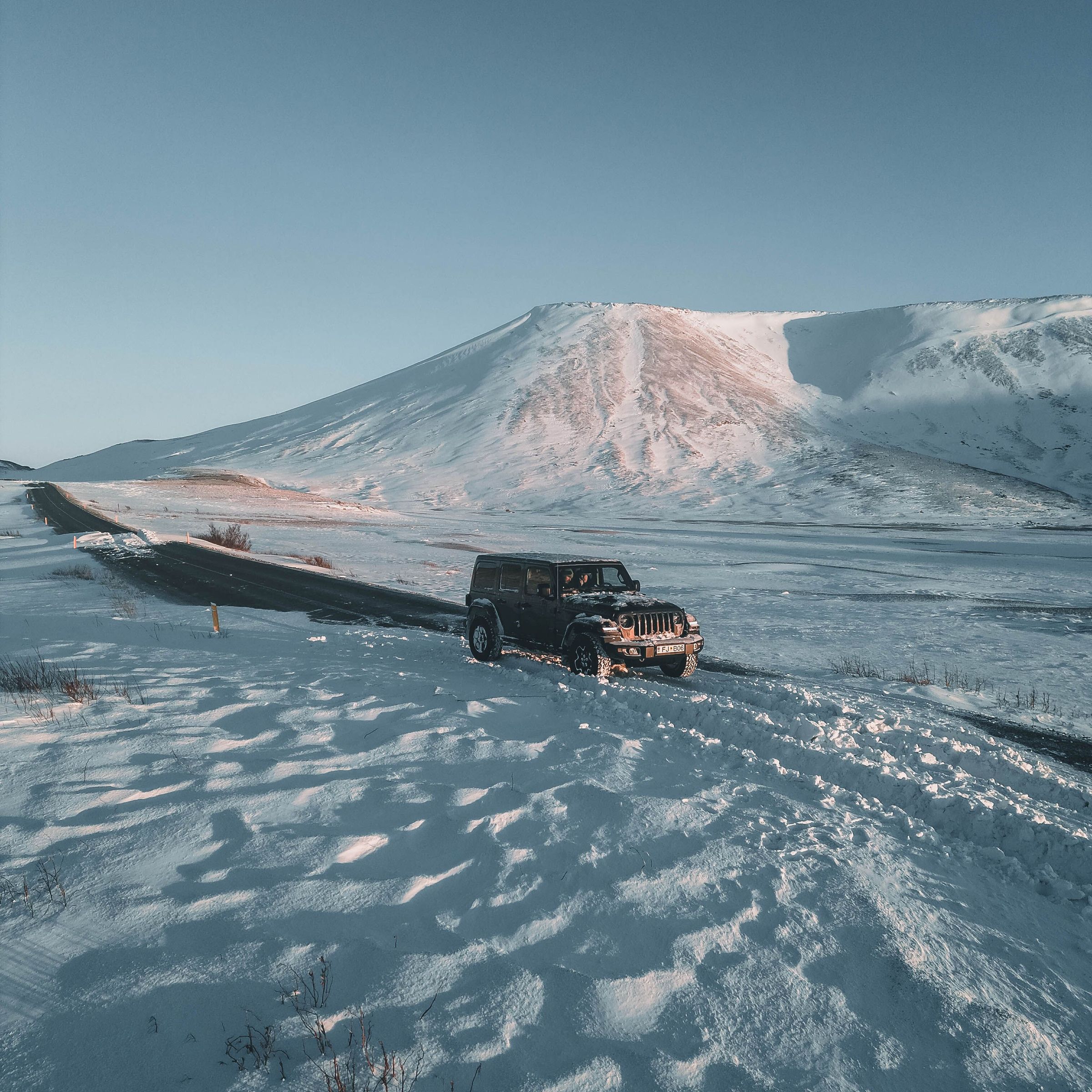 A dark SUV parked on a snowy road with snow-covered mountains under a clear blue sky.