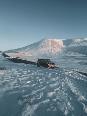A black Jeep Wrangler parked on a snowy shoulder beside a road, with a large snow-covered mountain under a clear blue sky.