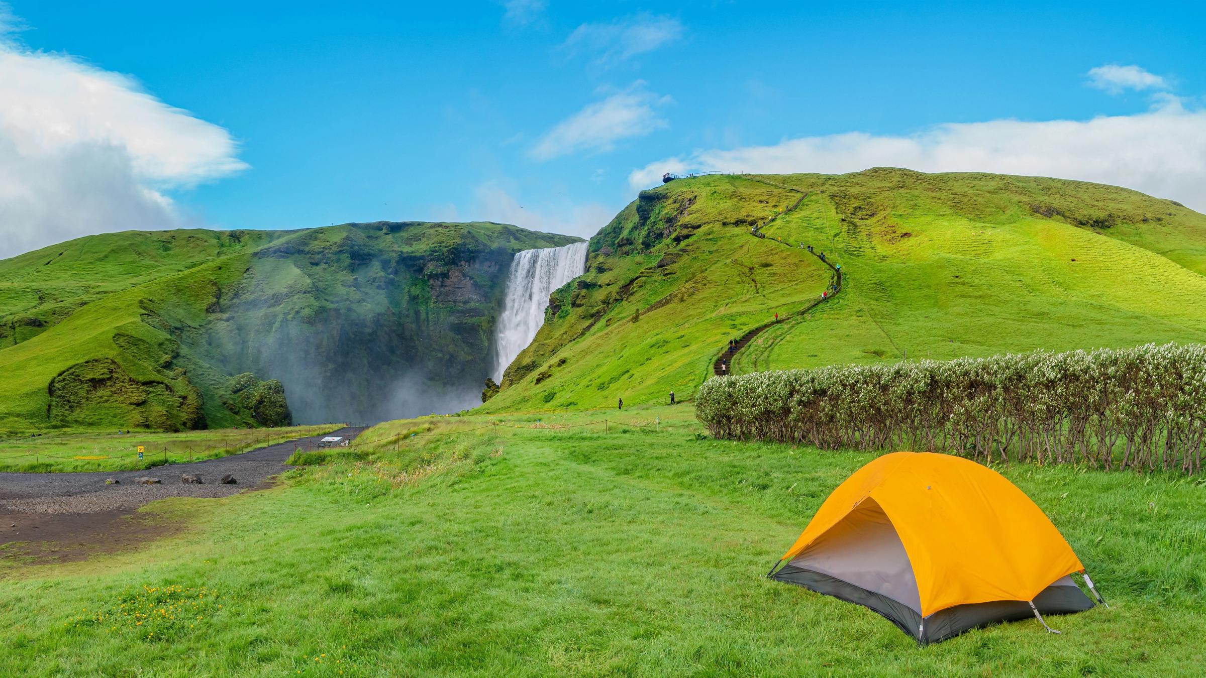 Panoramic view over camping site with orange tent, and tourists in front of famous Skogafoss waterfall, while hiking in Iceland, summer, scenic view.