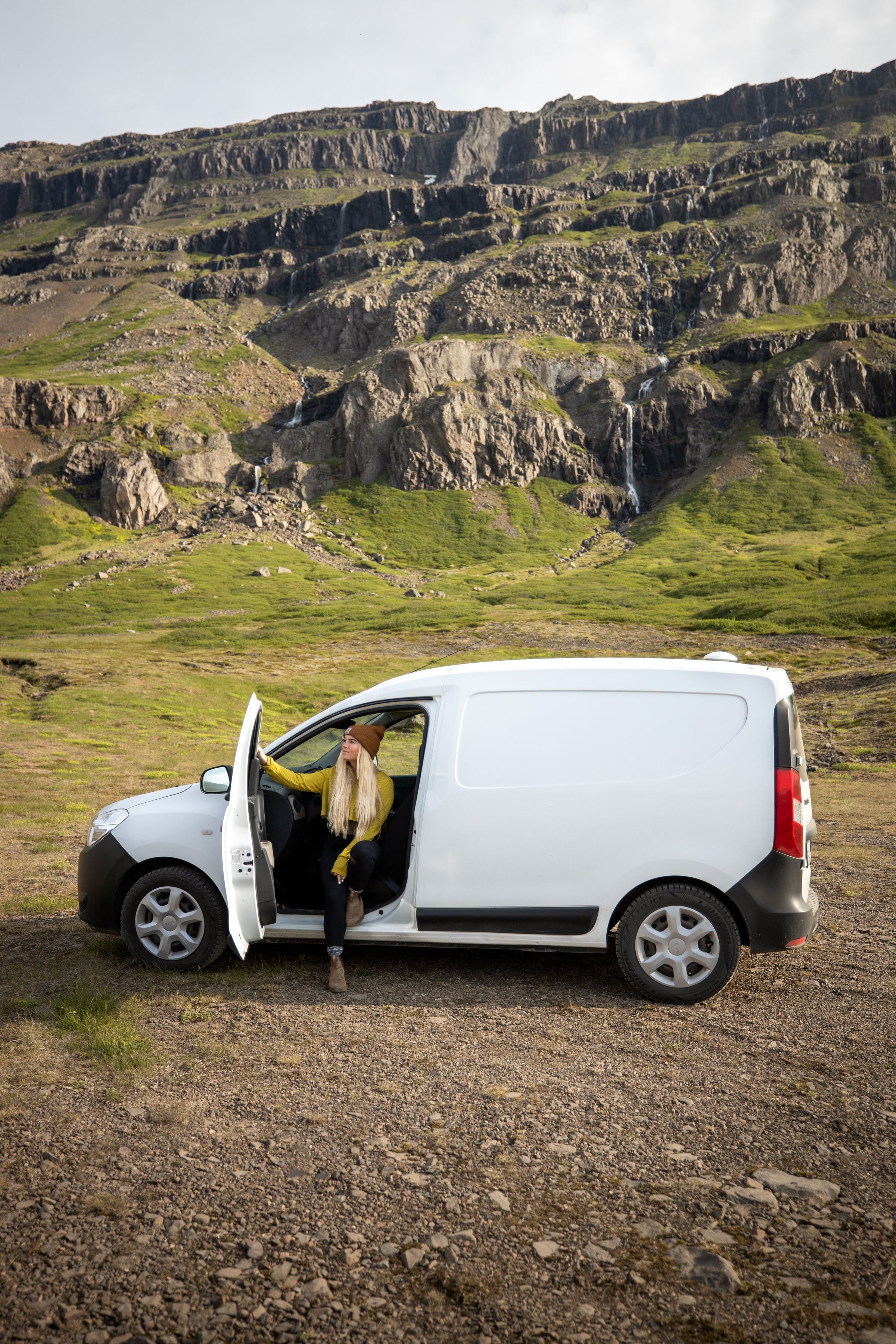 Seitenansicht eines kompakten, günstigen Campervans auf Schotter unterhalb von Klippen und einem Wasserfall, Island-Camper