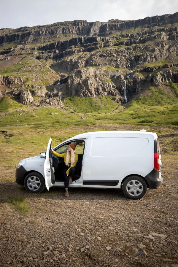 Seitenansicht eines kompakten, günstigen Campervans auf Schotter unterhalb von Klippen und einem Wasserfall, Island-Camper