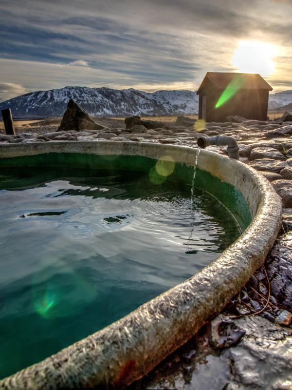 A circular concrete pool with green water, surrounded by rocks, a small wooden shack, and snow-capped mountains under a bright, low sun.