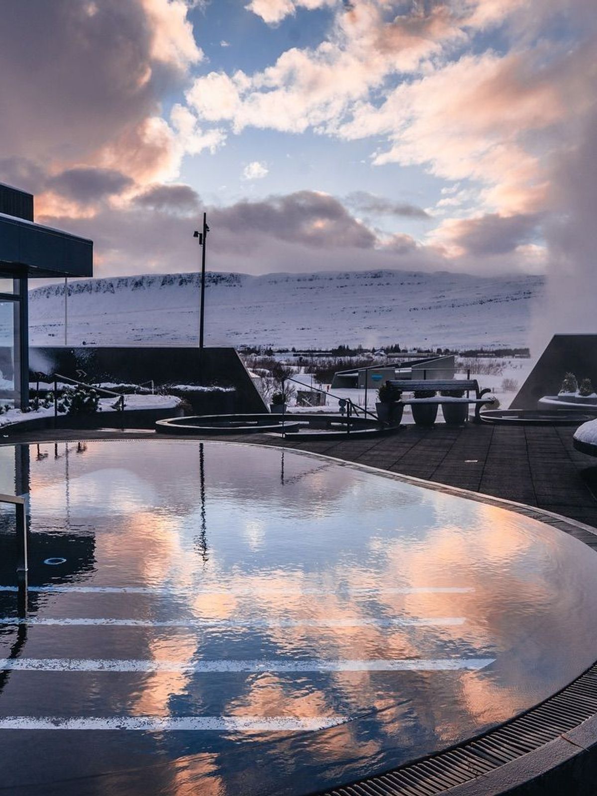 Outdoor thermal pool reflecting a colorful sky, with steam rising, modern buildings, and snow-covered mountains.