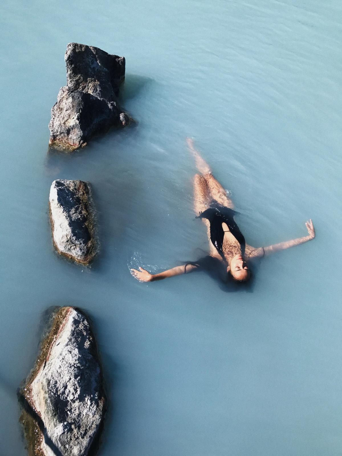 una mujer con un traje de baño negro está flotando boca arriba en un cuerpo de agua rodeado de rocas.