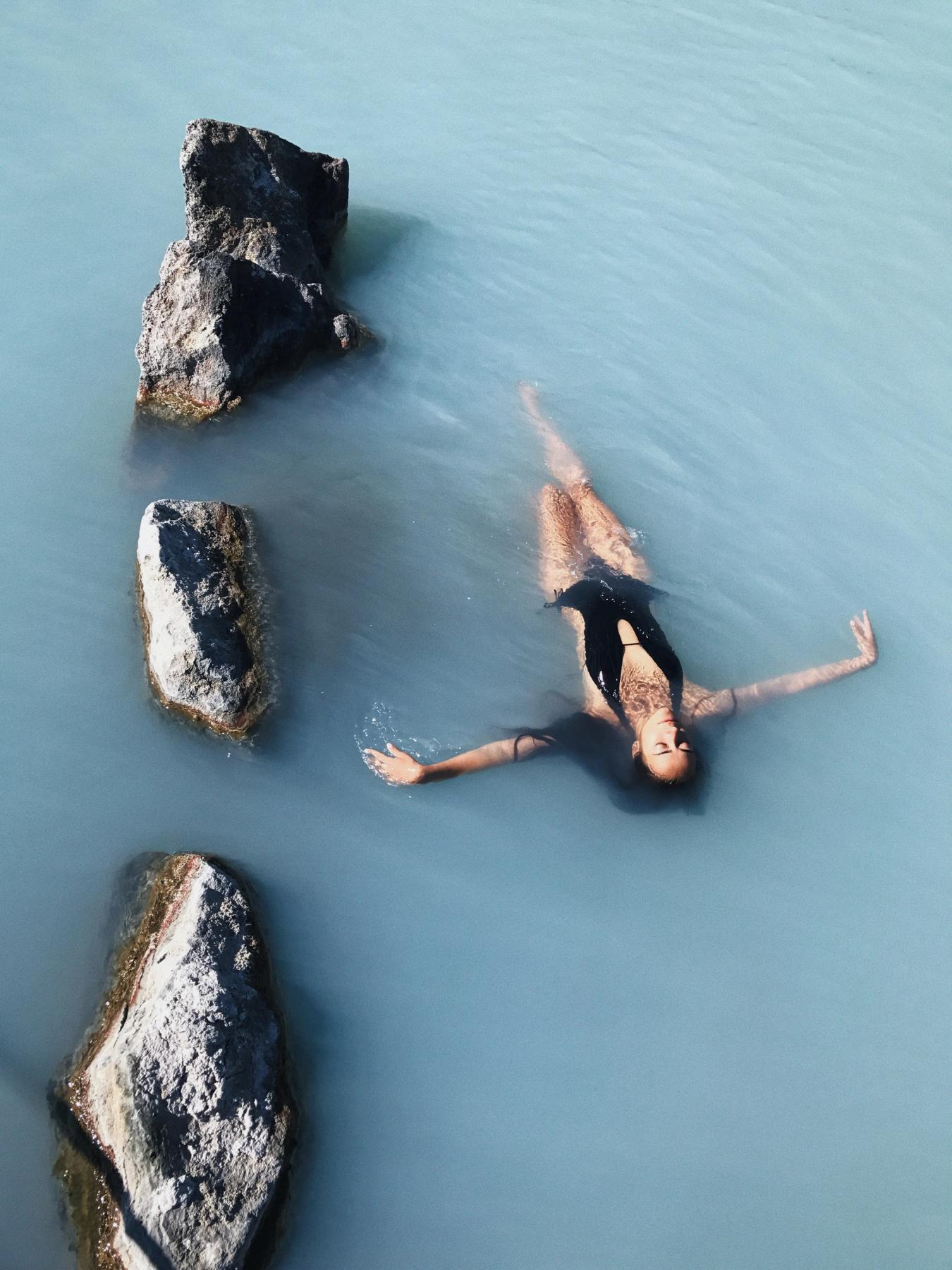 Woman floating peacefully in the milky blue geothermal waters of Sky Lagoon, surrounded by dark volcanic rocks.