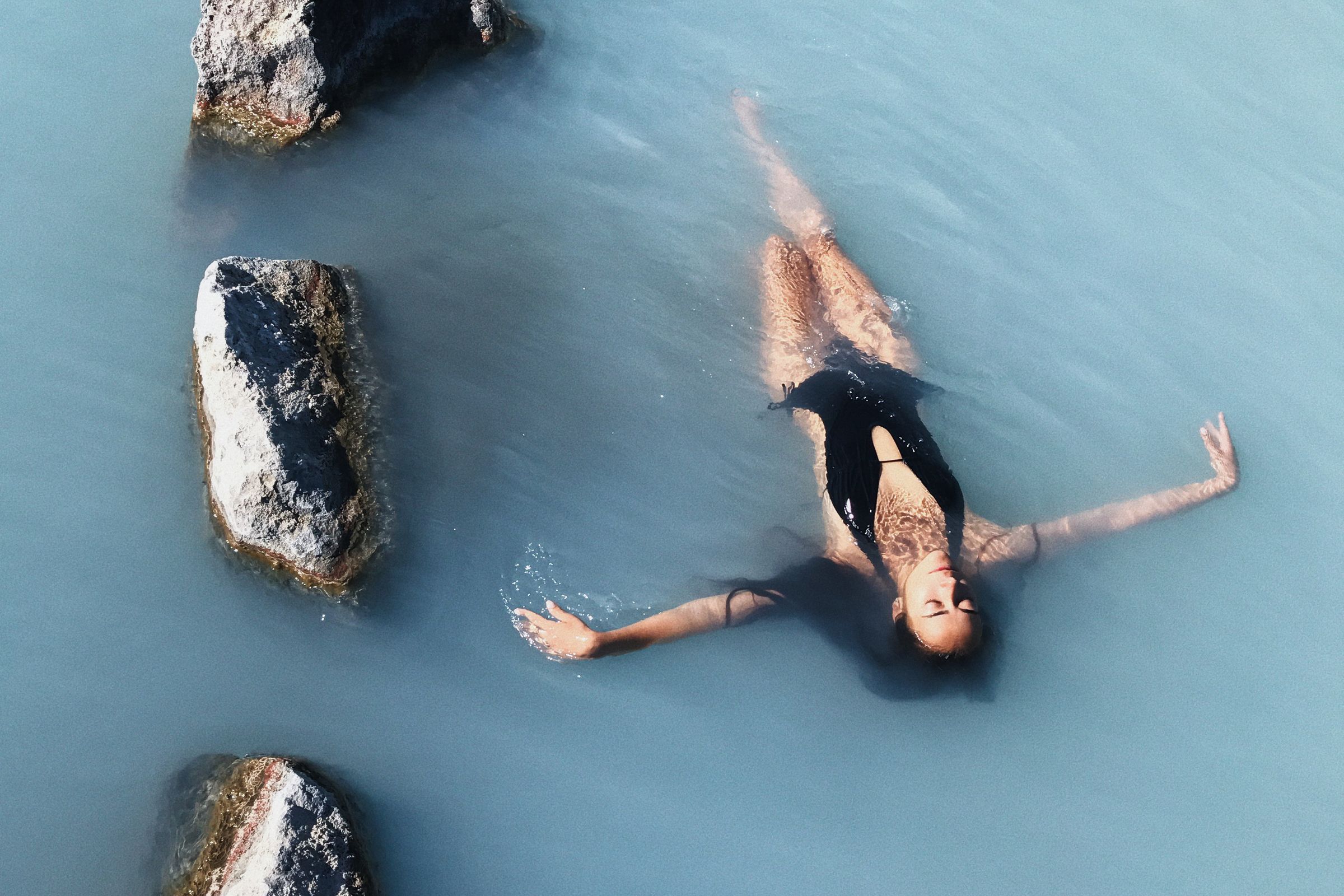 una mujer con un traje de baño negro está flotando boca arriba en un cuerpo de agua rodeado de rocas.