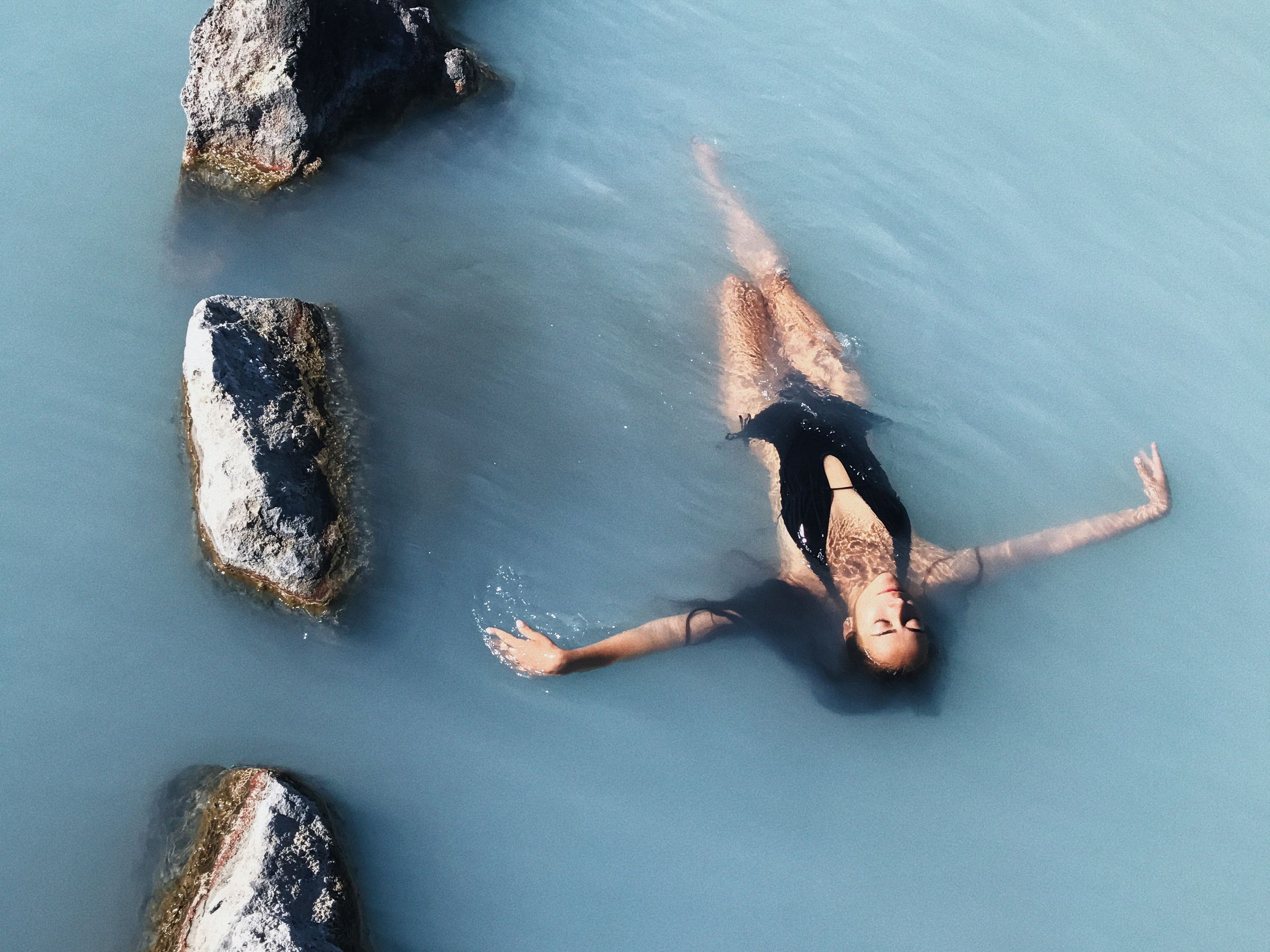 Woman floating peacefully in the milky blue geothermal waters of Sky Lagoon, surrounded by dark volcanic rocks.