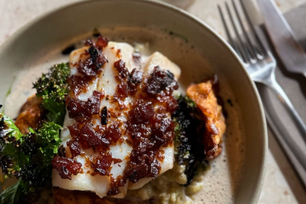 A plated meal of white fish topped with a dark relish, served with roasted potatoes, green vegetables, and a creamy sauce, with a glass of water and cutlery in the background.