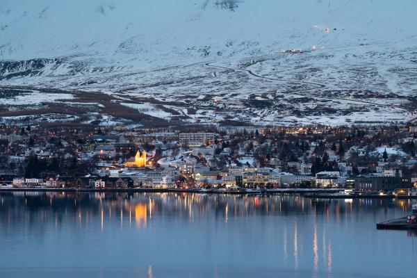 Una ciudad nevada al atardecer, con luces reflejándose en el agua tranquila y montañas en el fondo.