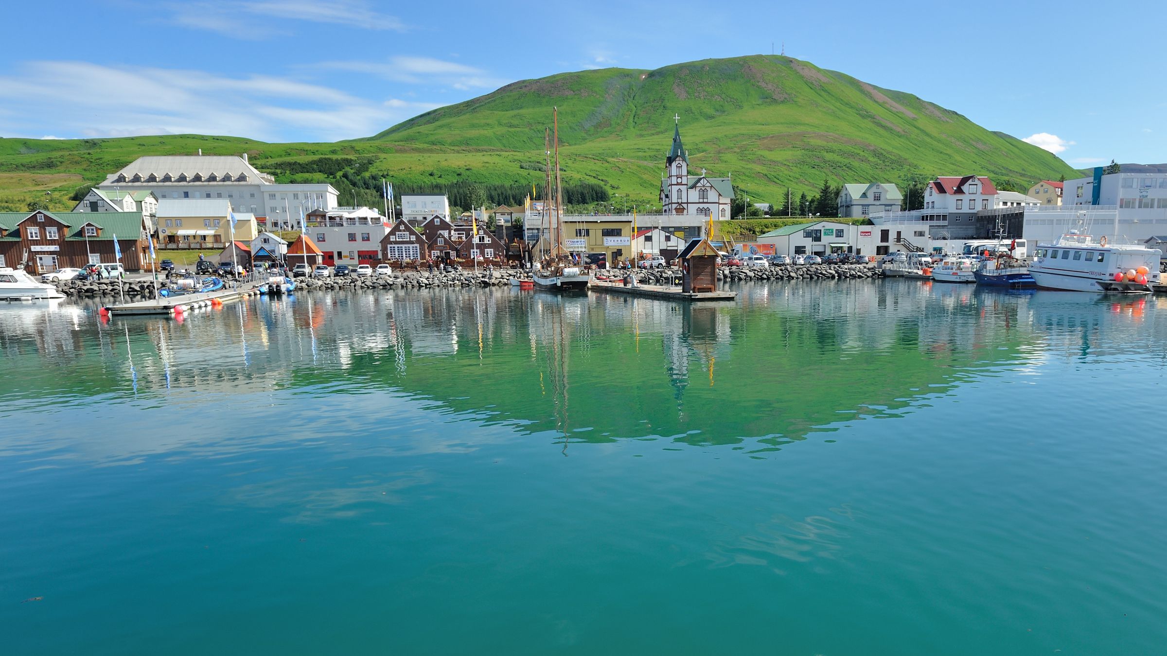 Colorful buildings and boats line a vibrant harbor with turquoise water, backed by a large green mountain.