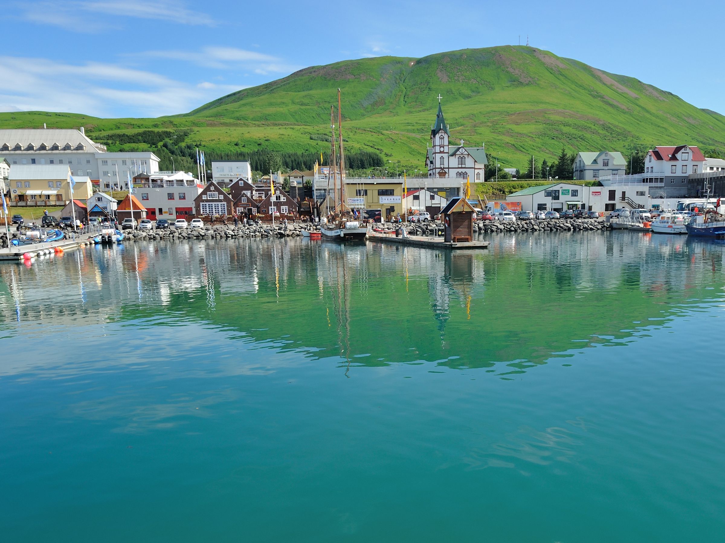 Colorful buildings and boats line a vibrant harbor with turquoise water, backed by a large green mountain.