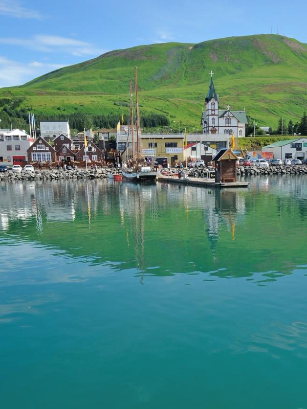 A colorful harbor town with a church steeple and boats docked in turquoise water, backed by a green mountain.