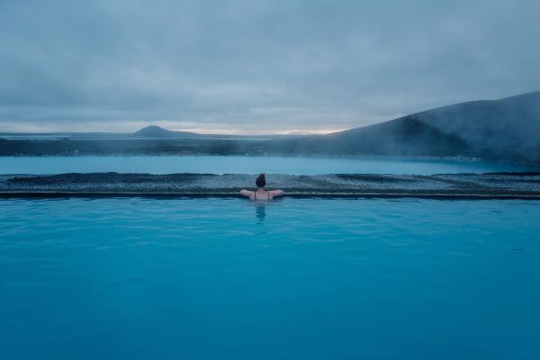 Person in a blue geothermal pool overlooking a misty, volcanic landscape.