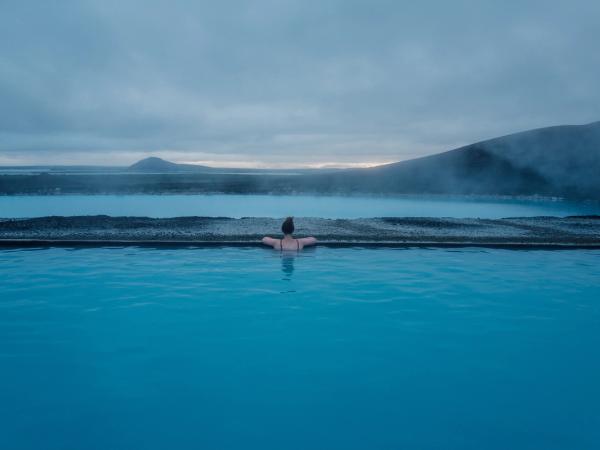 A person in a blue geothermal pool looking out at a misty blue lake and hills under a grey sky.