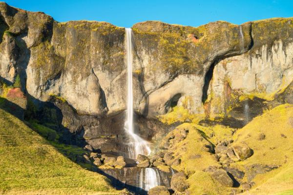 A narrow waterfall cascades down a mossy cliff face under a blue sky.