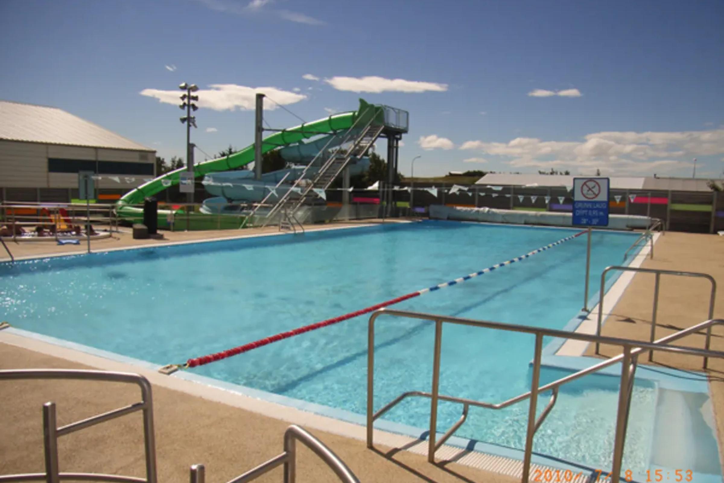 Una piscina al aire libre con un gran tobogán acuático verde y líneas de carril bajo un cielo azul.