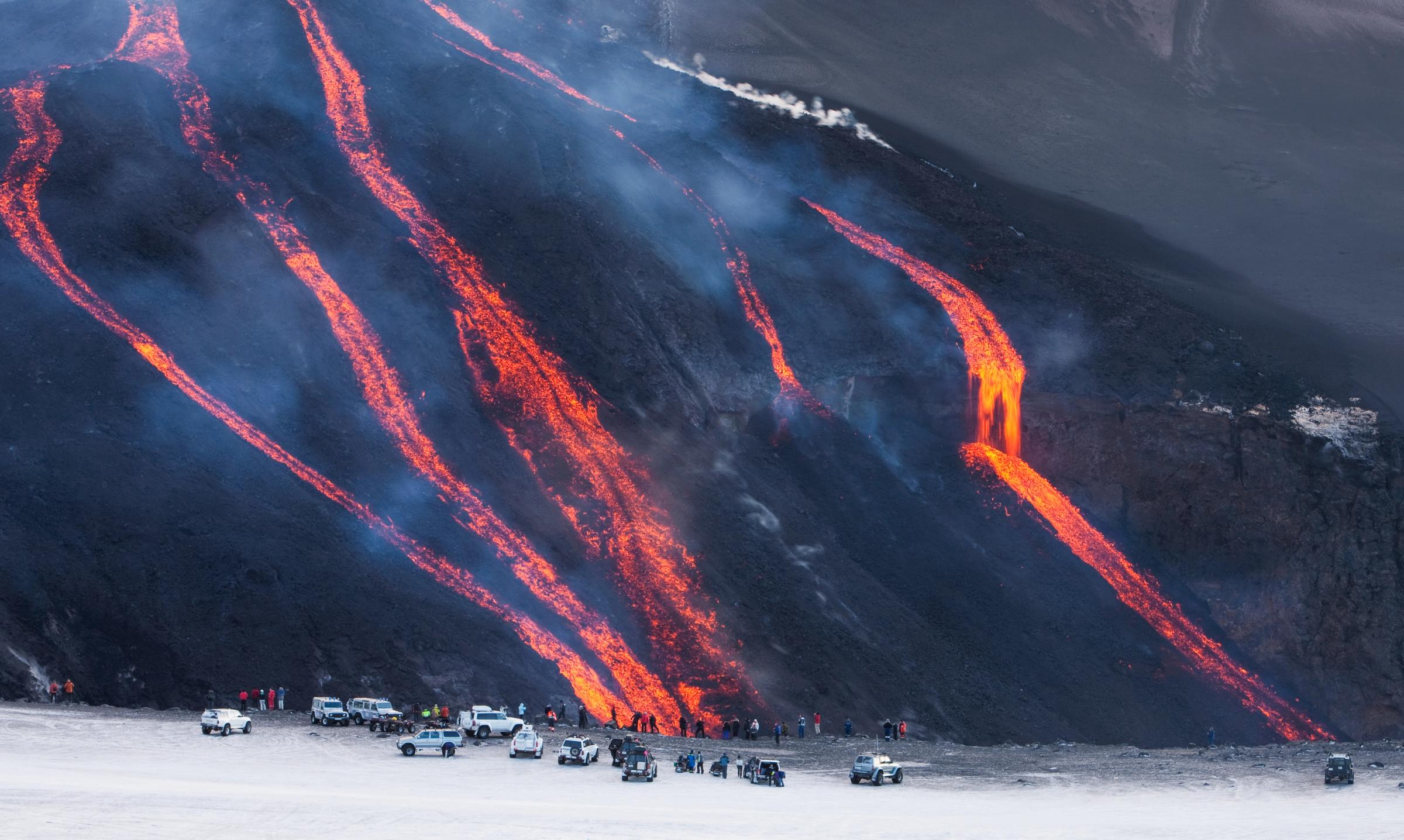Varios ríos de lava de un intenso color naranja fluyen por la pendiente oscura de un volcán, mientras personas y vehículos observan desde abajo.