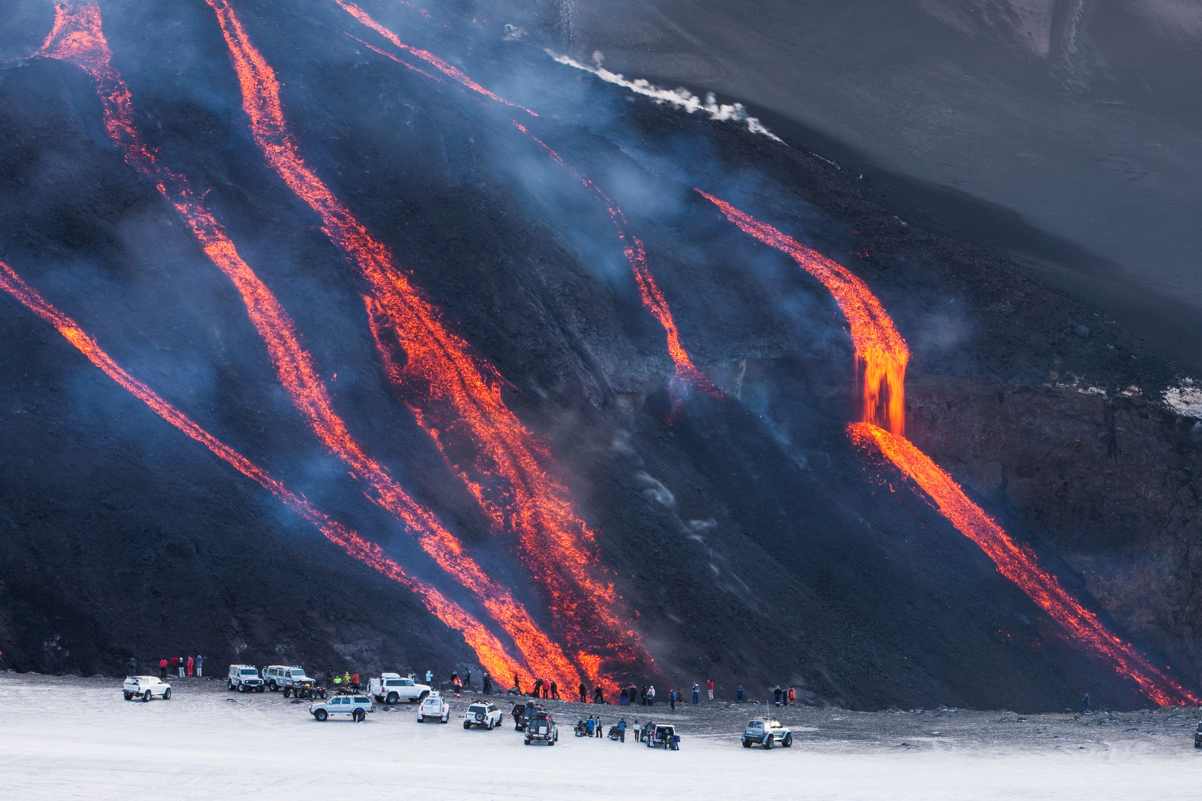 Varios ríos de lava de un intenso color naranja fluyen por la pendiente oscura de un volcán, mientras personas y vehículos observan desde abajo.