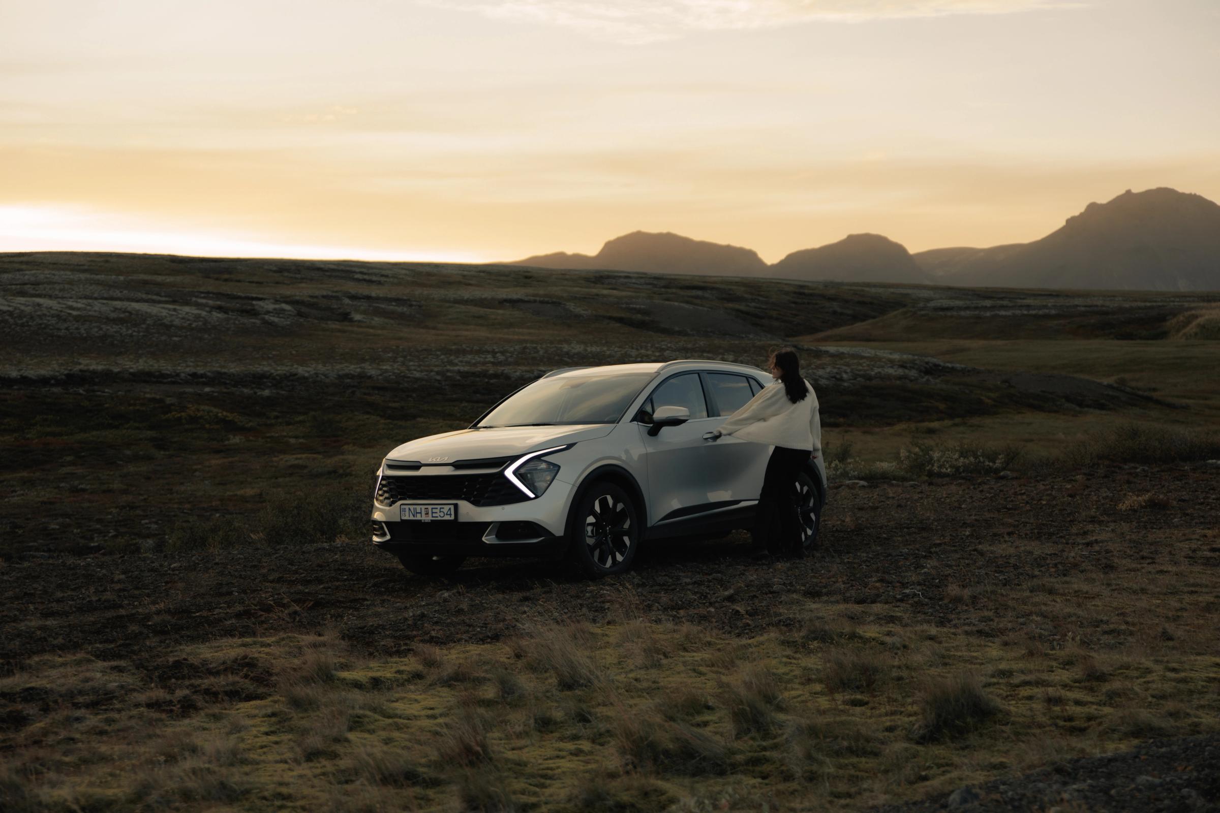 Kia Sportage sjálfkeyrslubíll á Íslandi, jeppi sem stendur í opnu hálendislandslagi með manneskju við hliðina