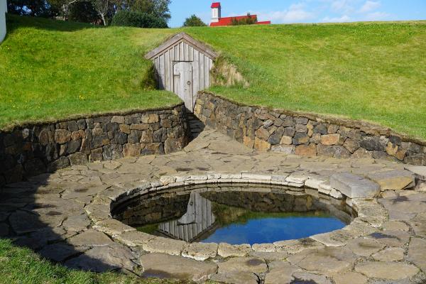 A circular stone hot spring pool with a wooden turf house built into a grassy hill behind it.