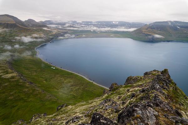 Fjord surrounded by green mountains under an overcast sky, with a road along the shore.