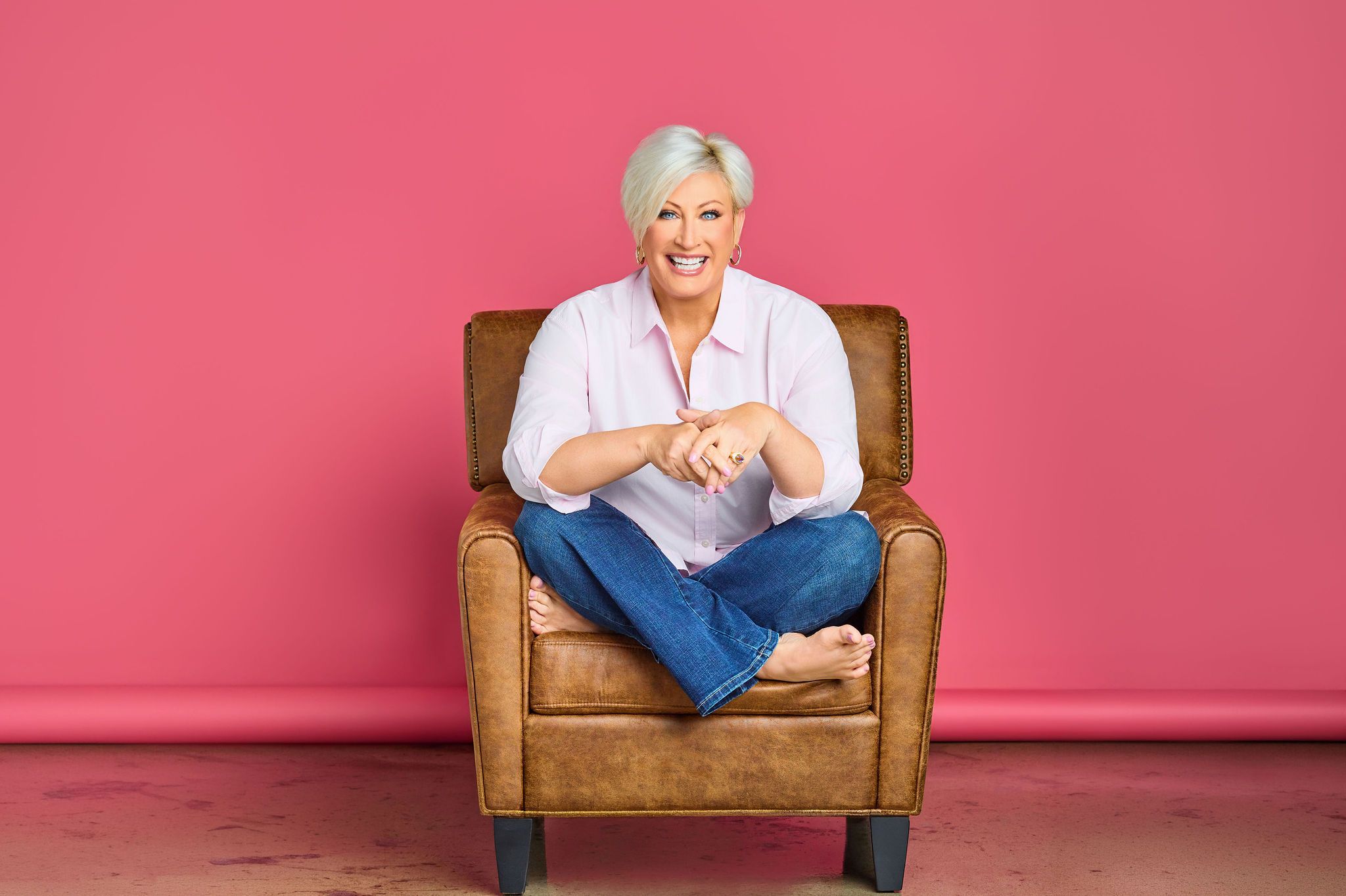Kim Gravel sitting cross-legged in a leather armchair on a pink backdrop
