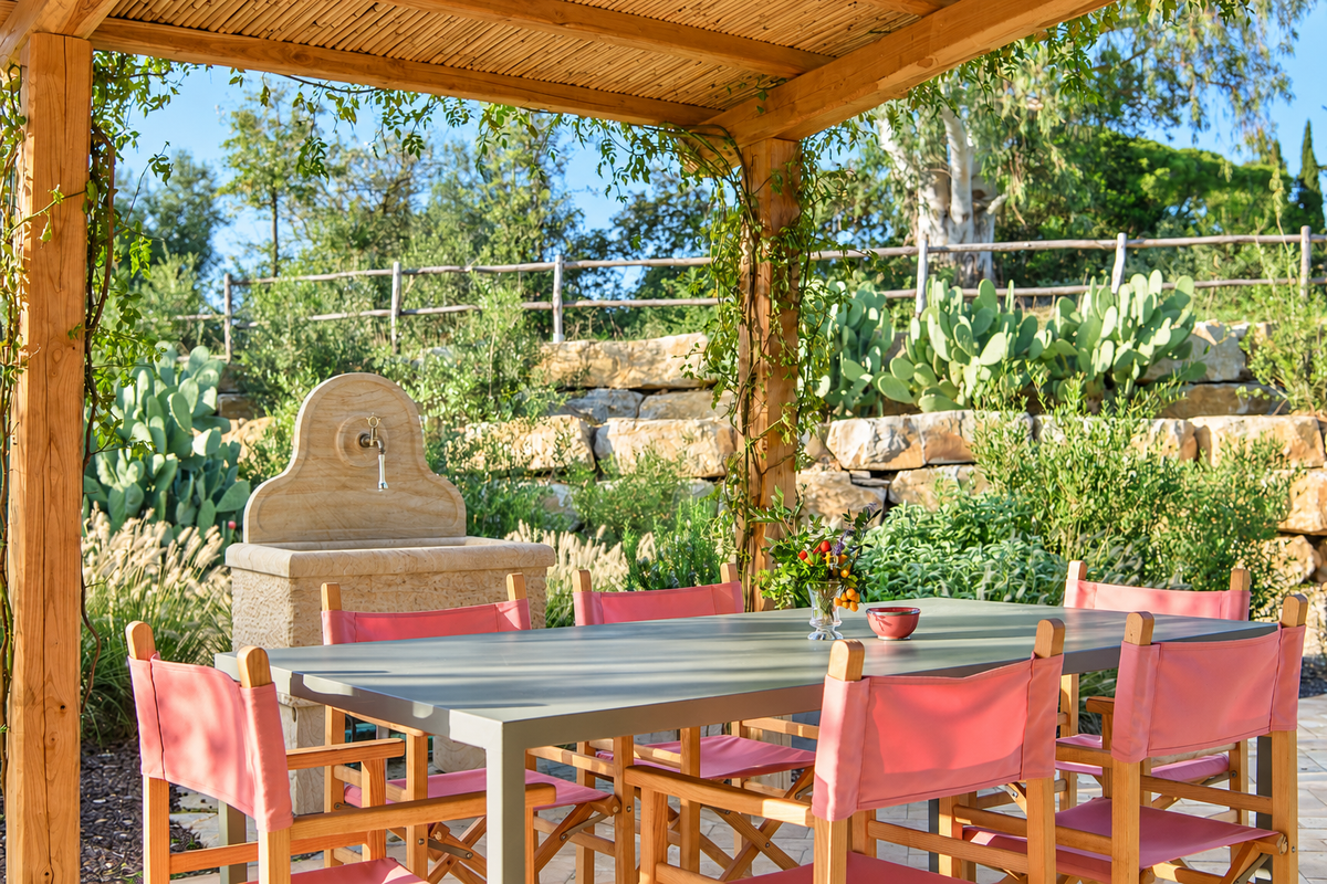 Lunch table with pink chairs under a cane pergola, beside a stone fountain