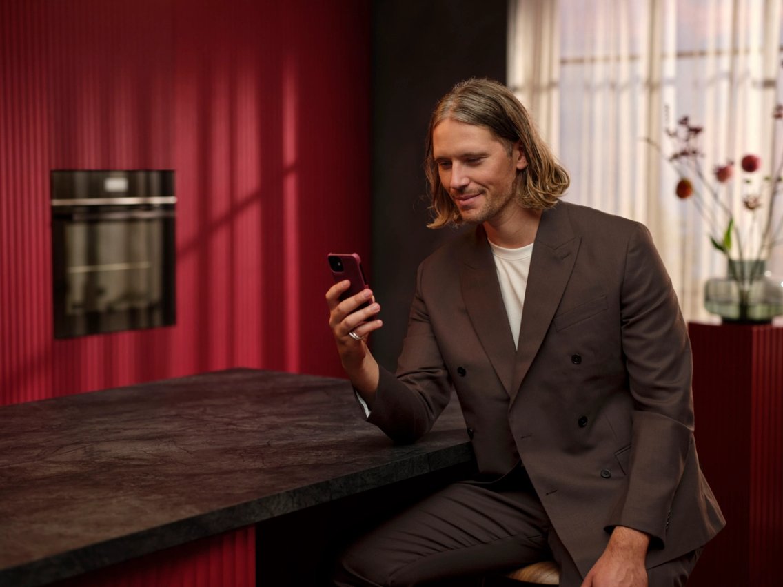 Man in a red kitchen smiling at his smartphone, with a built-in AEG oven behind him