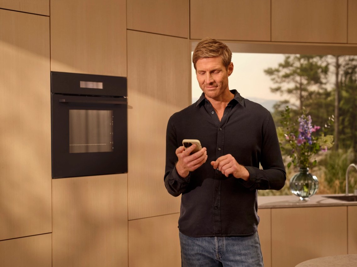 Man in a light wood kitchen looking at his smartphone, with a built-in Electrolux oven beside him