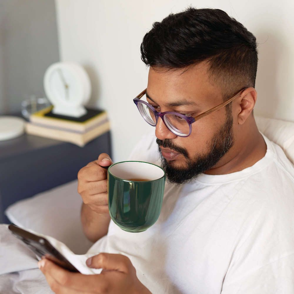 Man Drinking Coffee in Bed Looking at His Phone