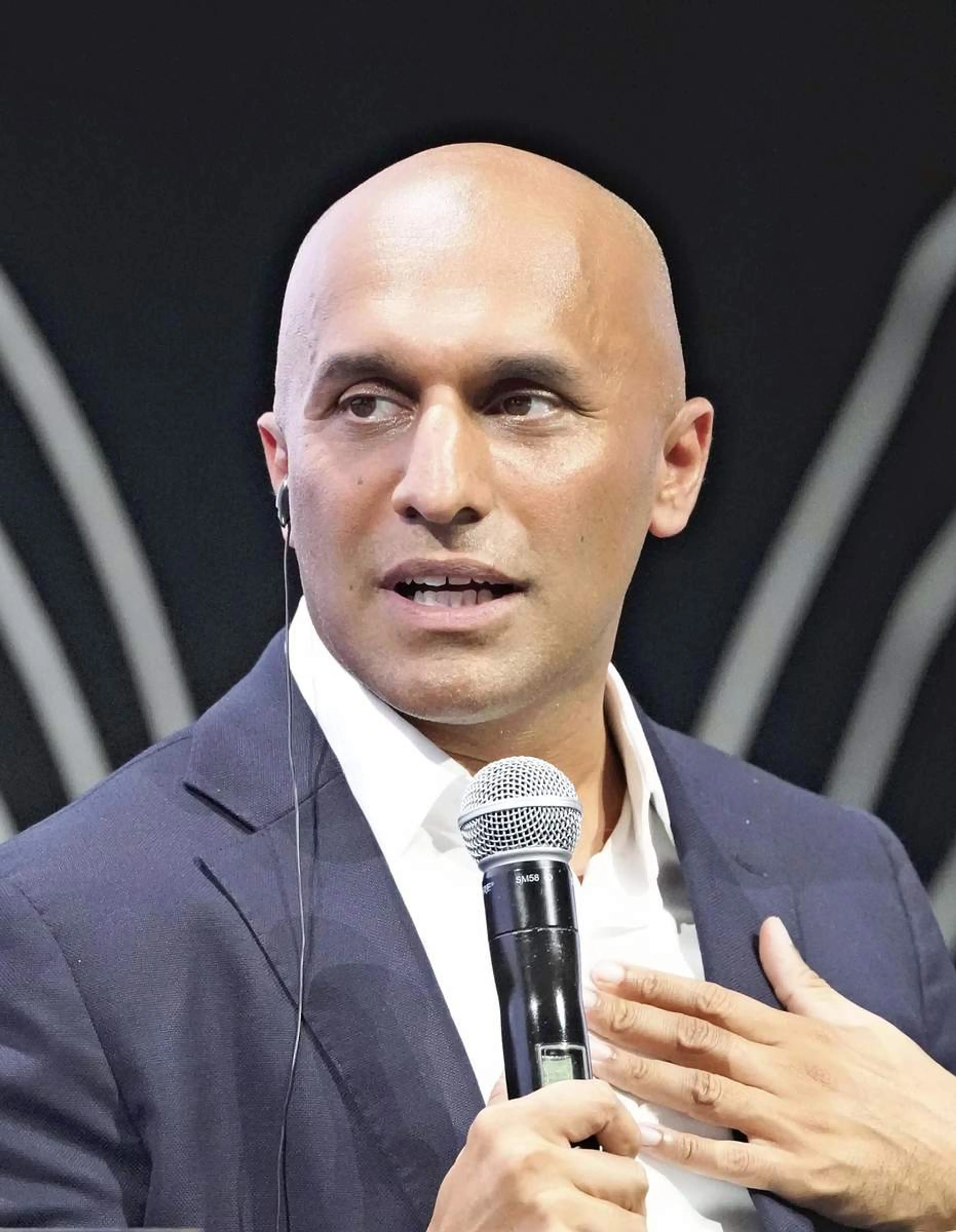 Qasar Younis speaking, holding a microphone and gesturing with his right hand, wearing a navy blazer and open-collar white shirt