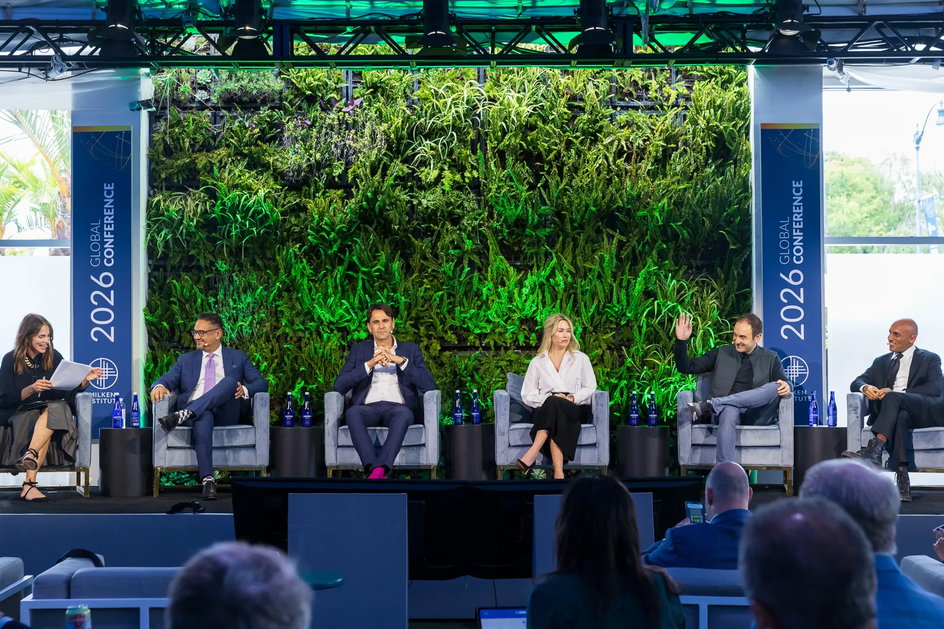 Six panelists seated in gray chairs on a conference stage at the 2026 Milken Institute Global Conference, with a large living plant wall behind them and an audience visible in the foreground.