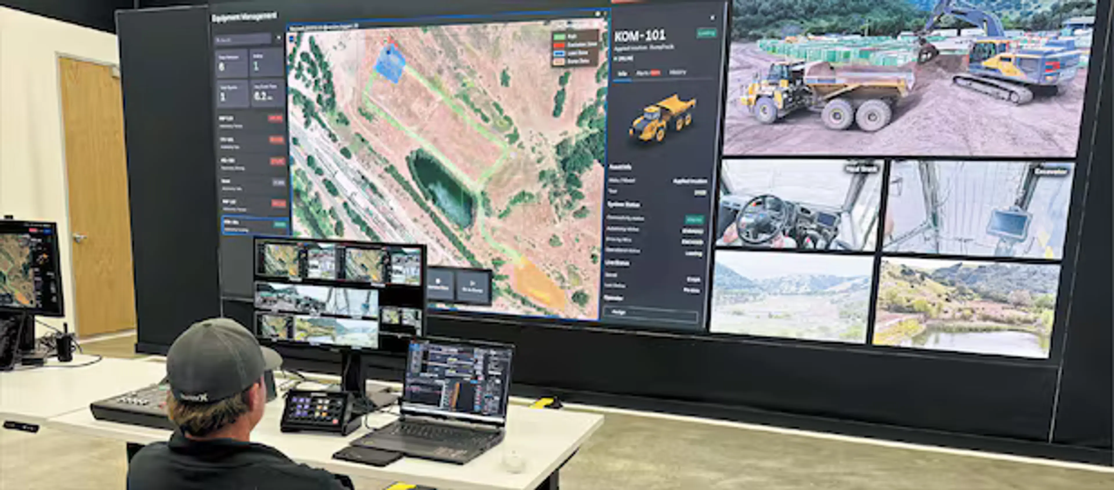 Here's my suggested alt text, with a few options depending on the context: Concise (for decorative/editorial use):  Operator at a control station monitoring a large display wall showing an equipment management dashboard with aerial maps and live camera feeds of mining or construction vehicles