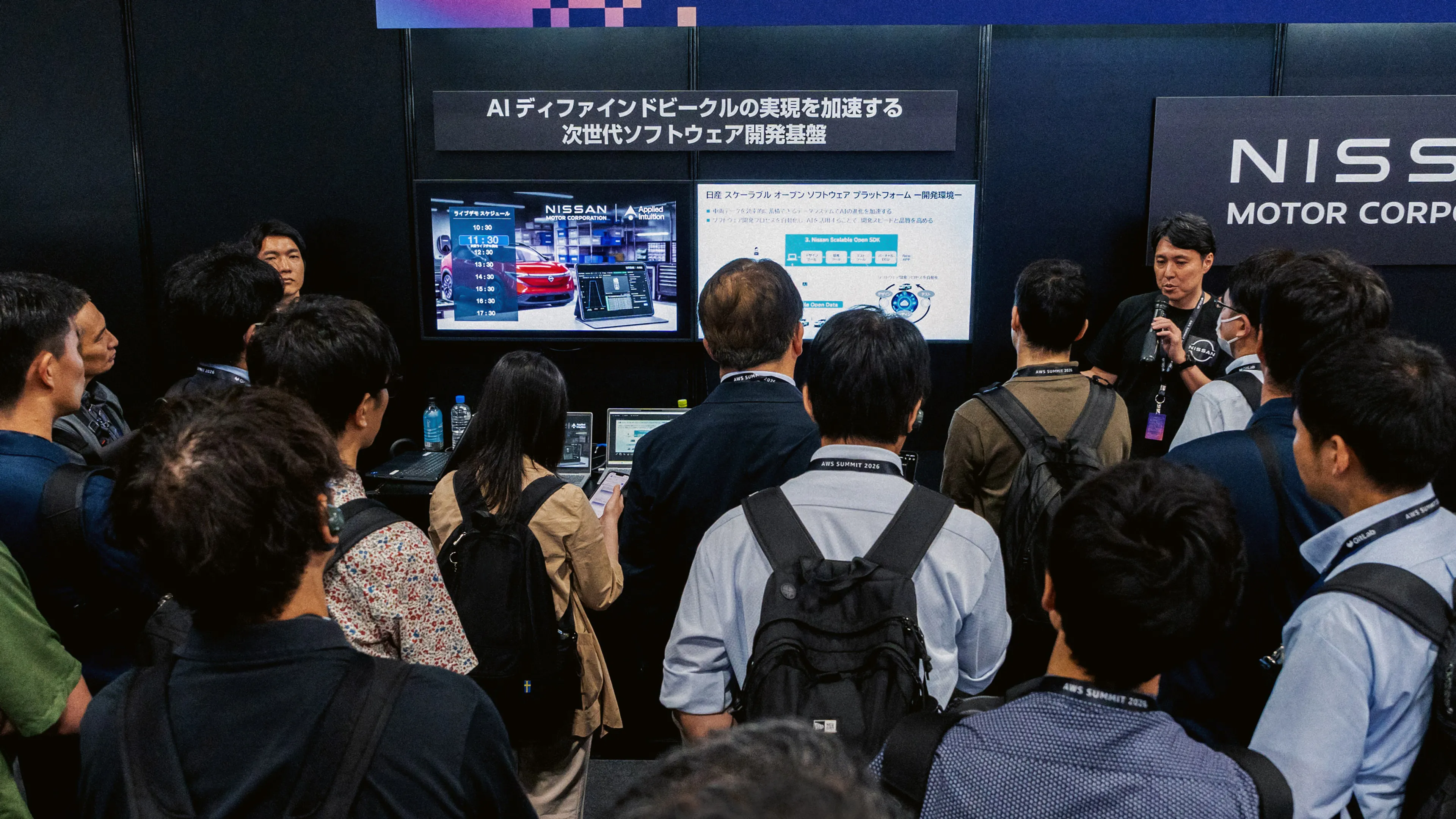 A presenter speaks to a crowd of attendees at a trade show booth.