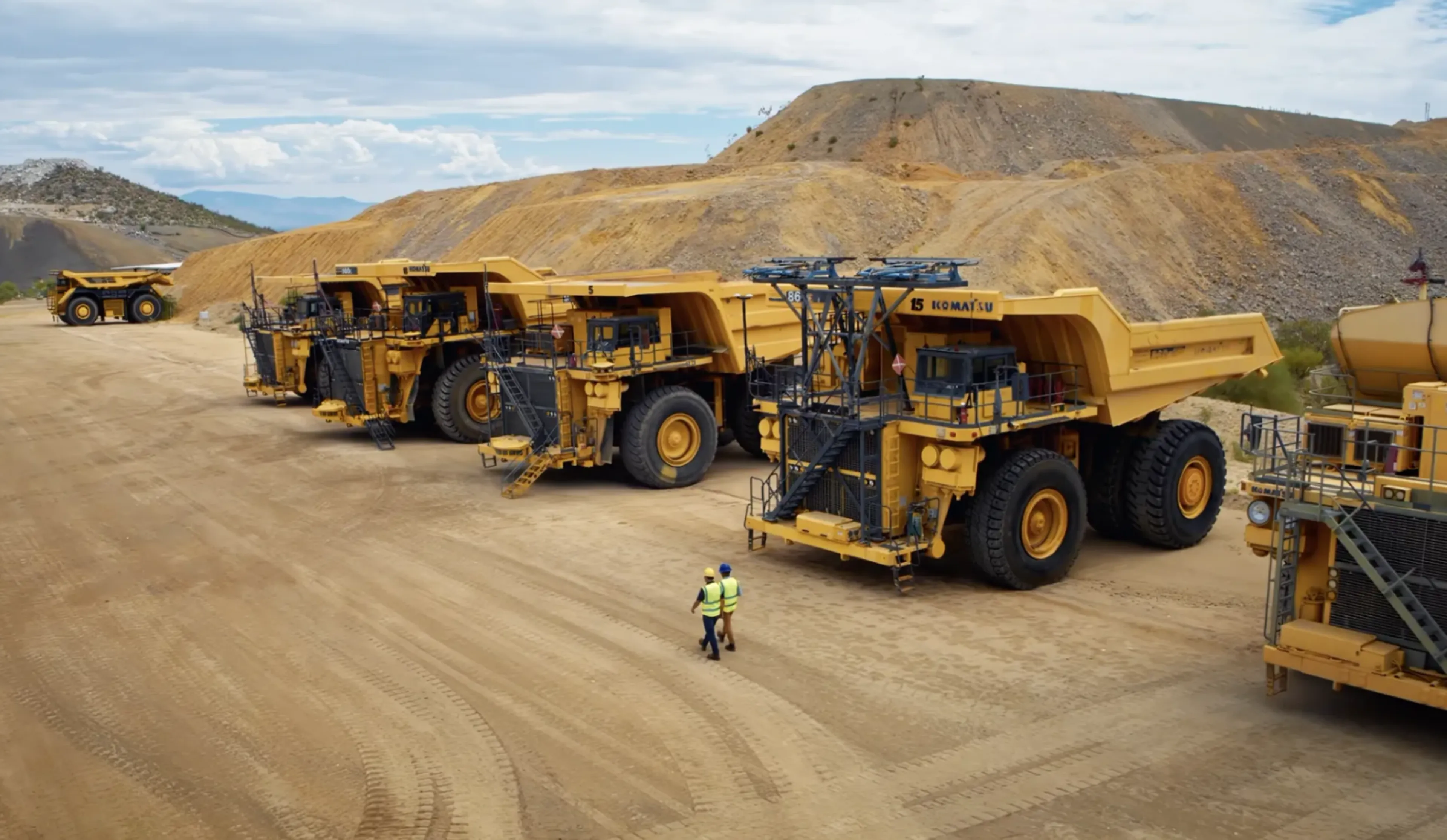 Row of large mining haul trucks at an open-pit mine, with two workers in high-visibility gear walking nearby.