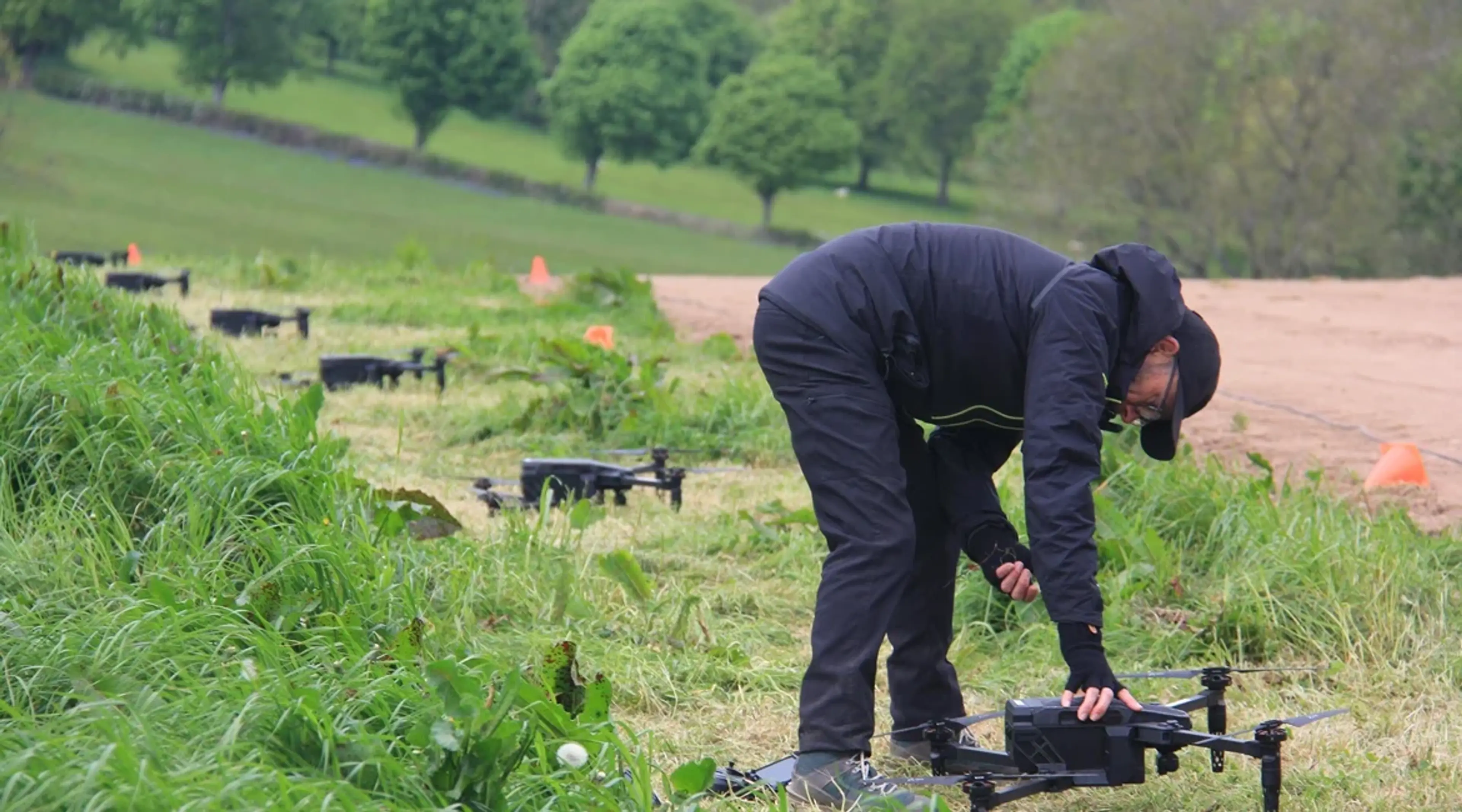 Person in dark outdoor clothing bends over to adjust a drone in a grassy field, with several other drones positioned nearby
