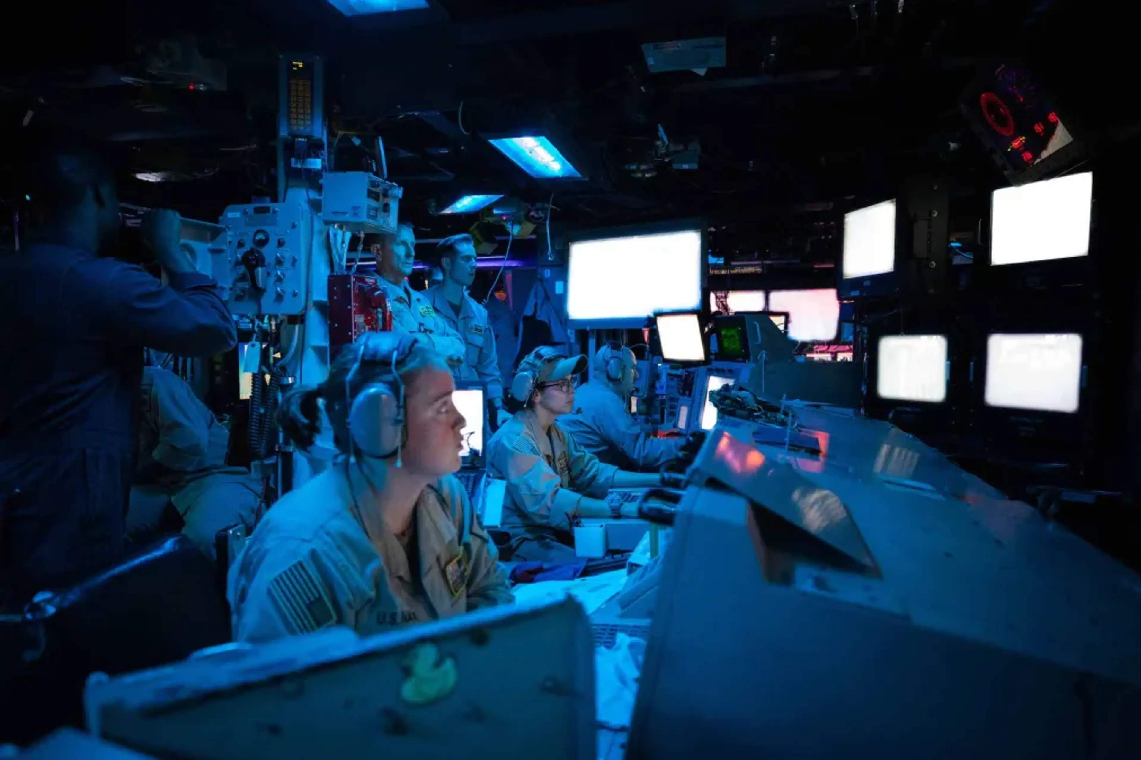 U.S. Navy sailors in military uniforms and headsets operate multiple computer workstations and monitors in a darkened combat information center, illuminated by blue ambient lighting