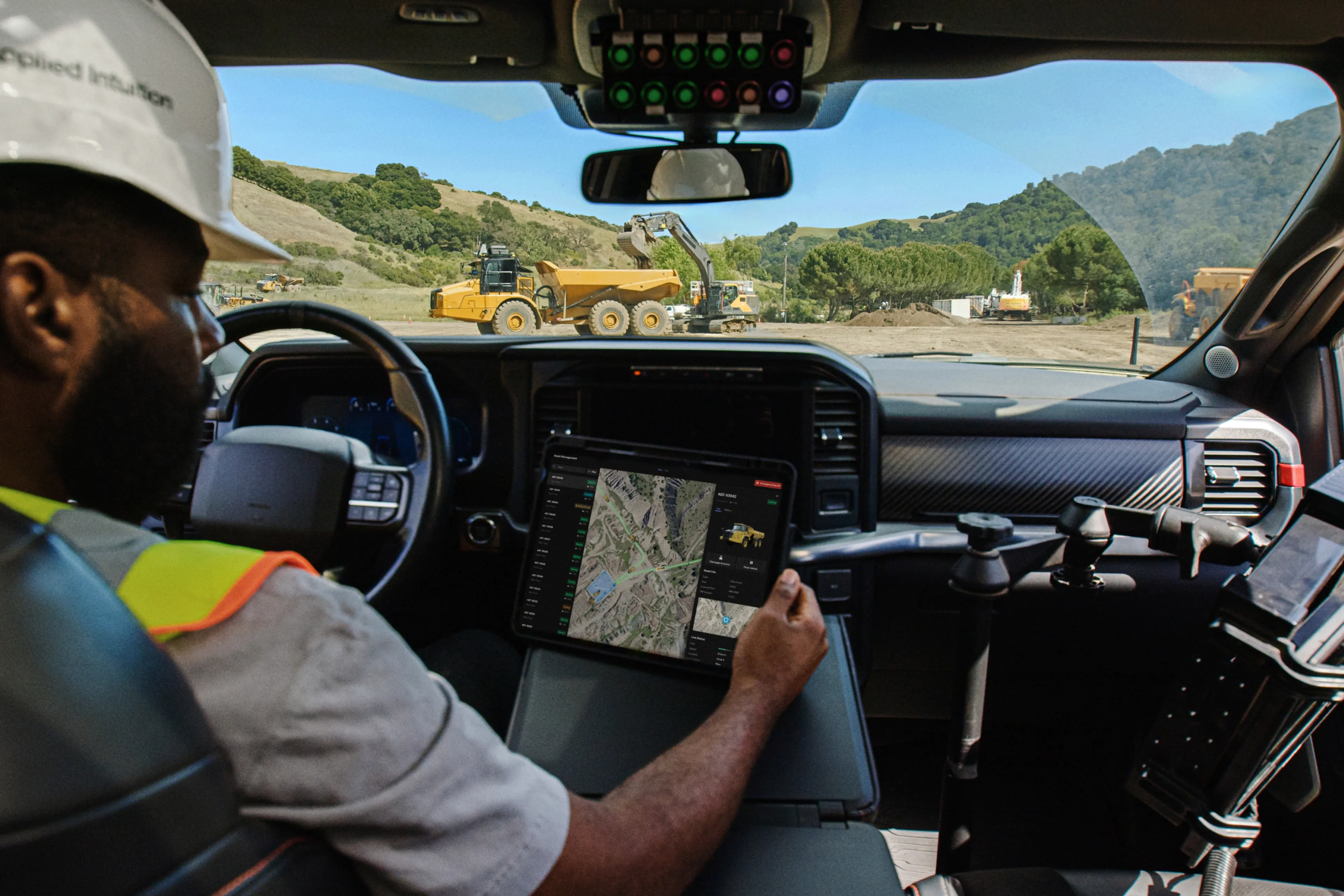 A construction worker sitting in a vehicle cab reviews a tablet showing a fleet management map, with heavy construction equipment visible through the windshield