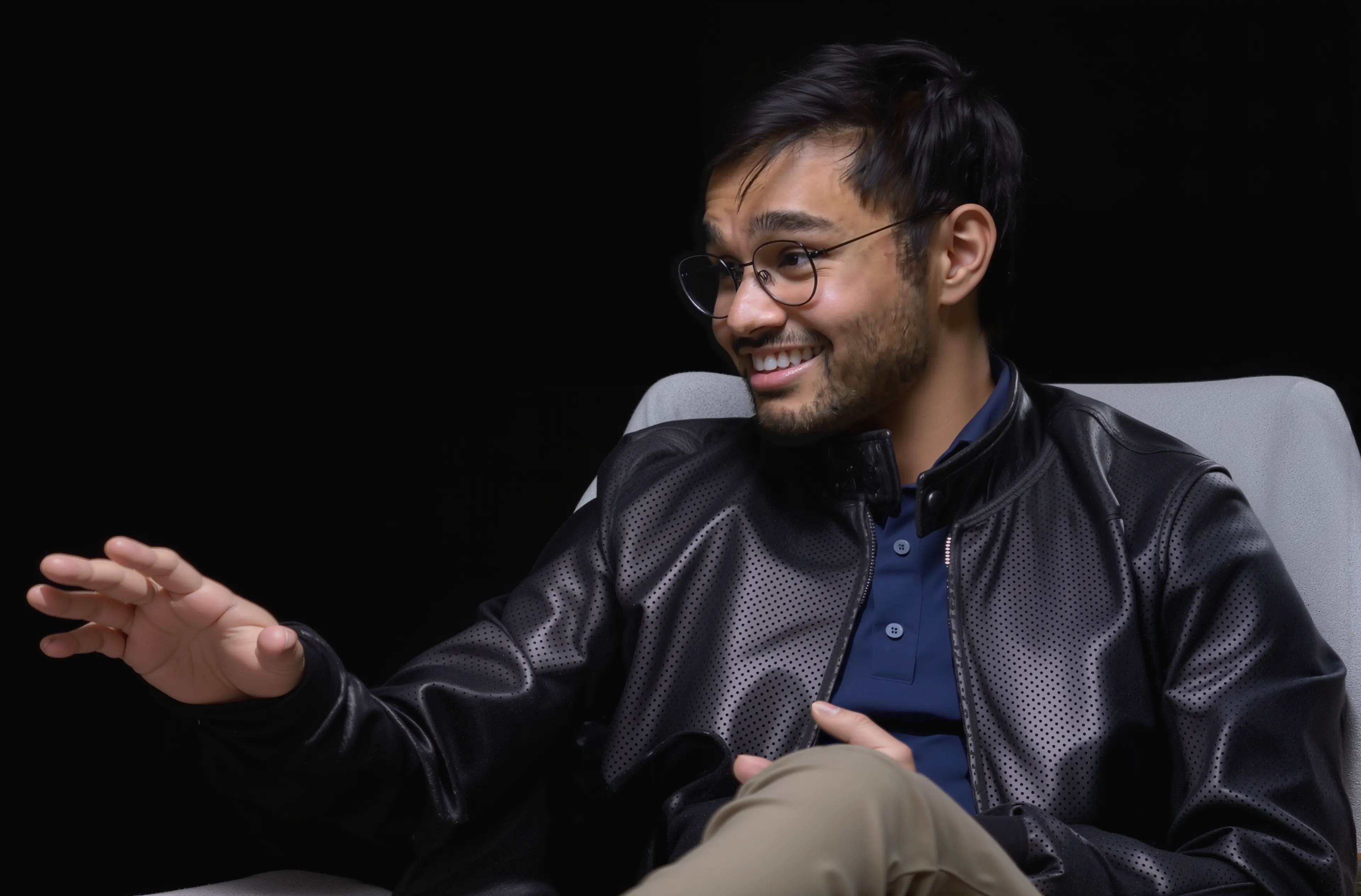 Malhar Patel smiles and gestures with one hand while seated in a gray chair against a black background.
