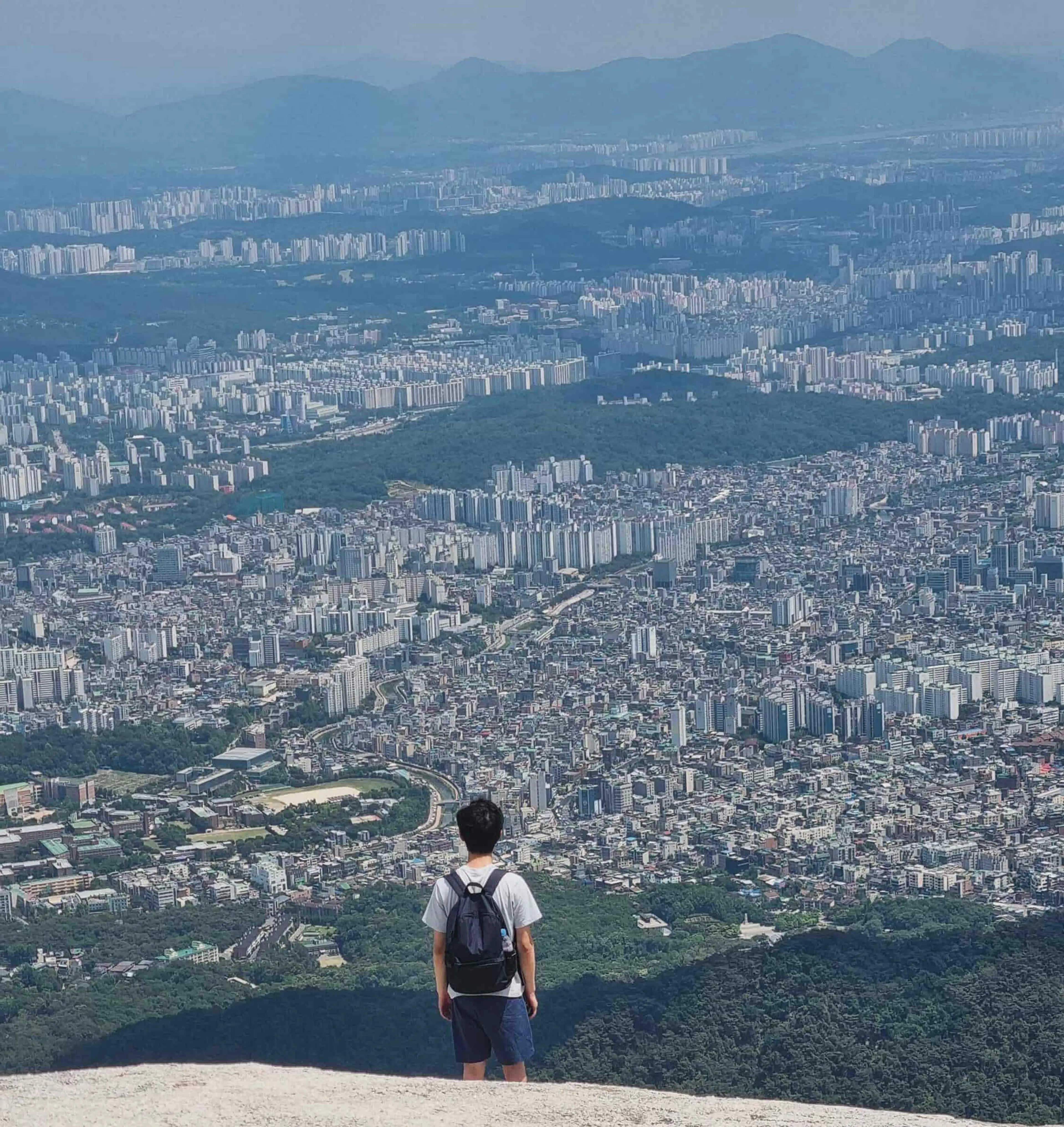 Rear view of person wearing a backpack as they are atop a mountain looking over Seoul, Korea