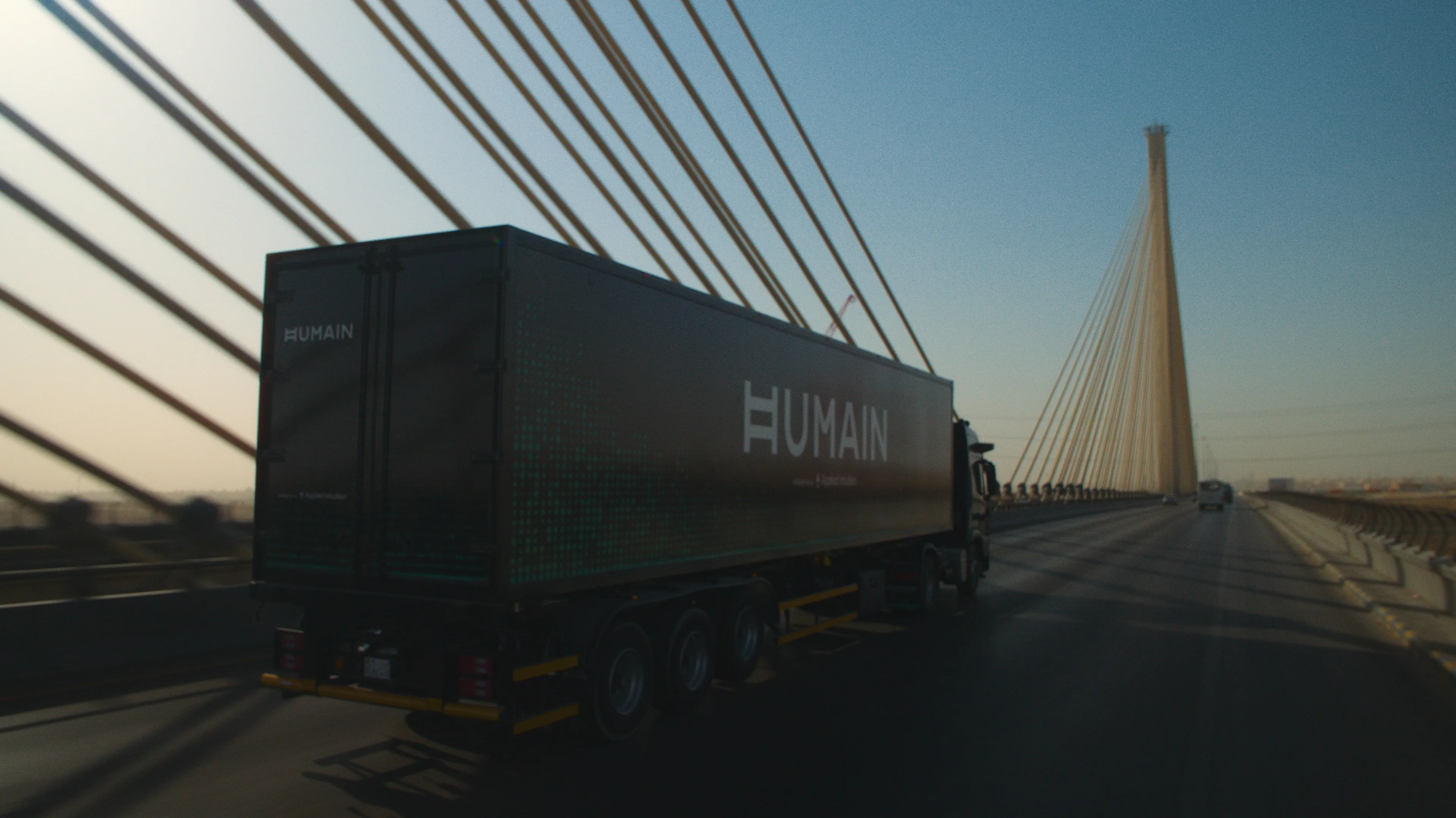 Rear view of a black HUMAIN semi-truck and trailer driving across a long bridge in low evening light, with cables fanning out from a tall pylon ahead.