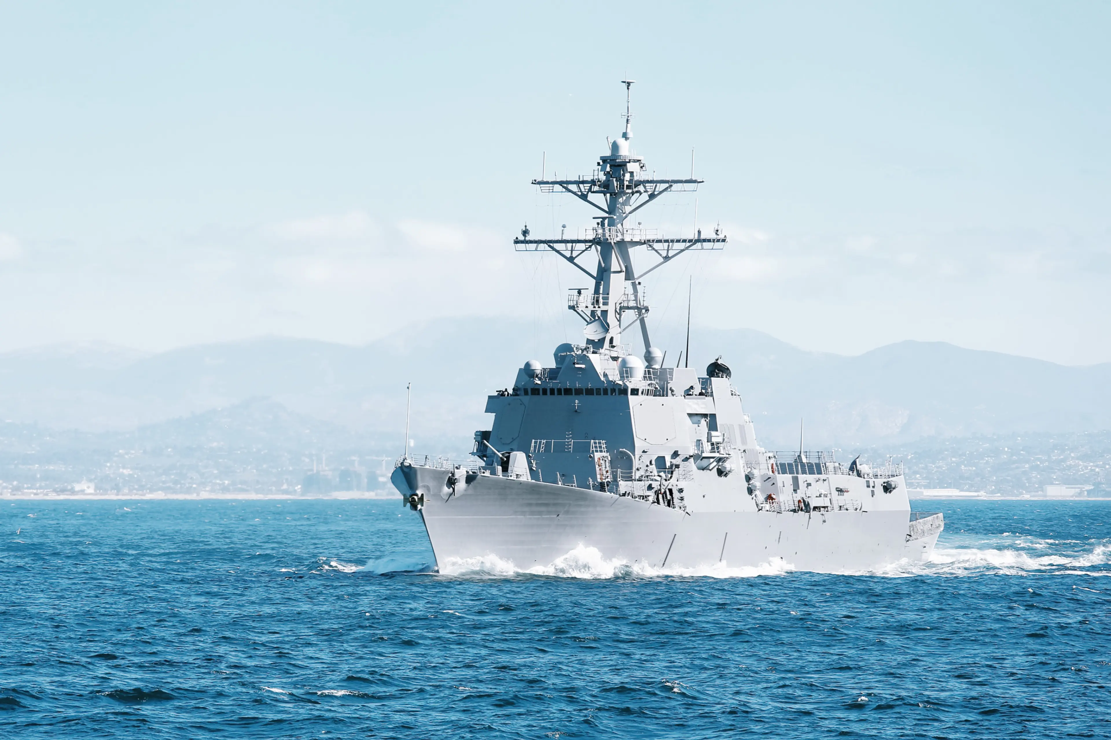 Naval destroyer cutting through rough ocean waves, overlaid with soft, colorful light flares in pink, purple, and rainbow hues against a gray sky