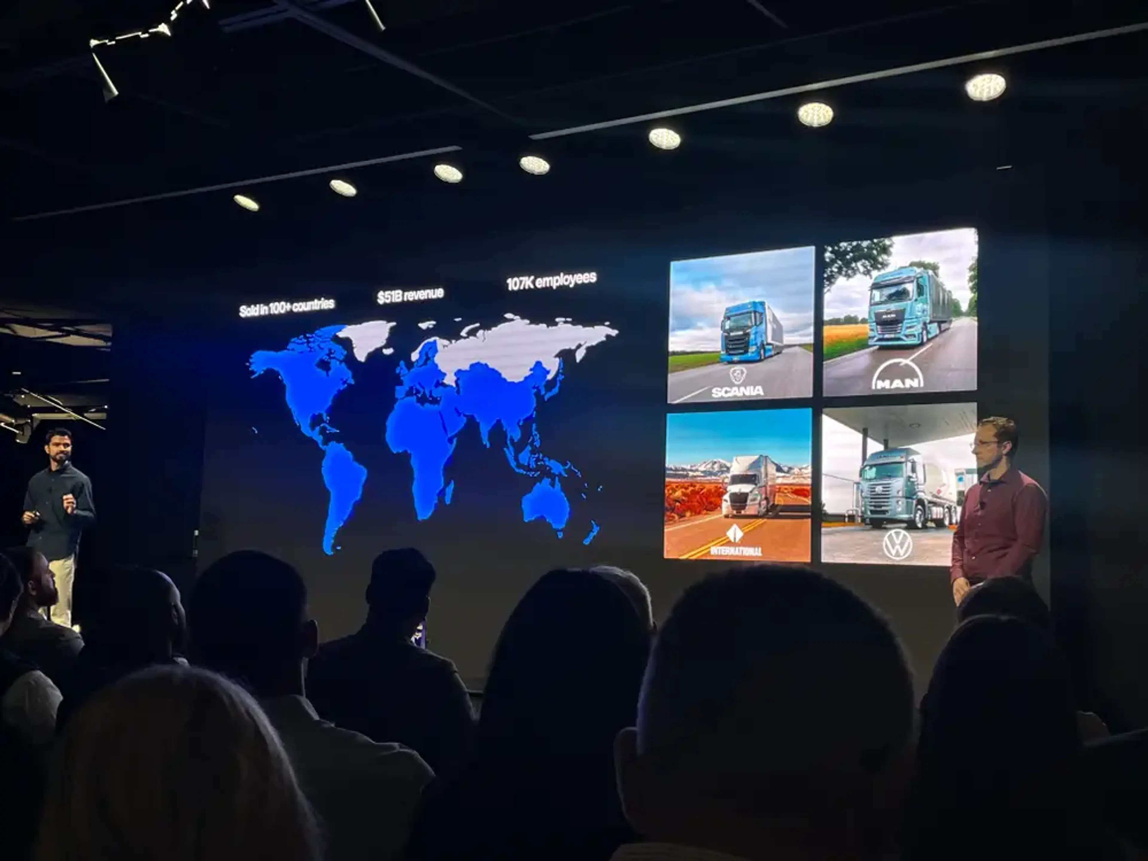 Here's suggested alt text for this image: Short version (for decorative/contextual use):  "Presentation slide showing a world map and truck brand logos including Scania, MAN, International, and Volkswagen, with stats: sold in 100+ countries, $51B revenue, 107K employees